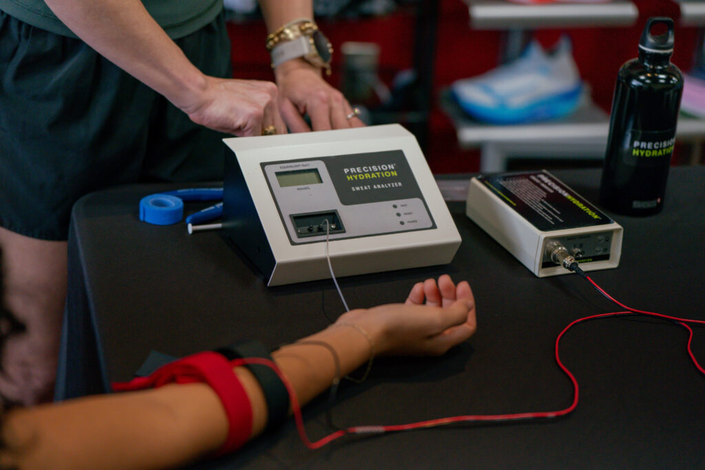 A person’s arm with electrodes attached rests on a table next to a Precision Hydration sweat analyzer device for a sweat test for athletes, while Jena Brown stands nearby. A water bottle and tape are also on the table.