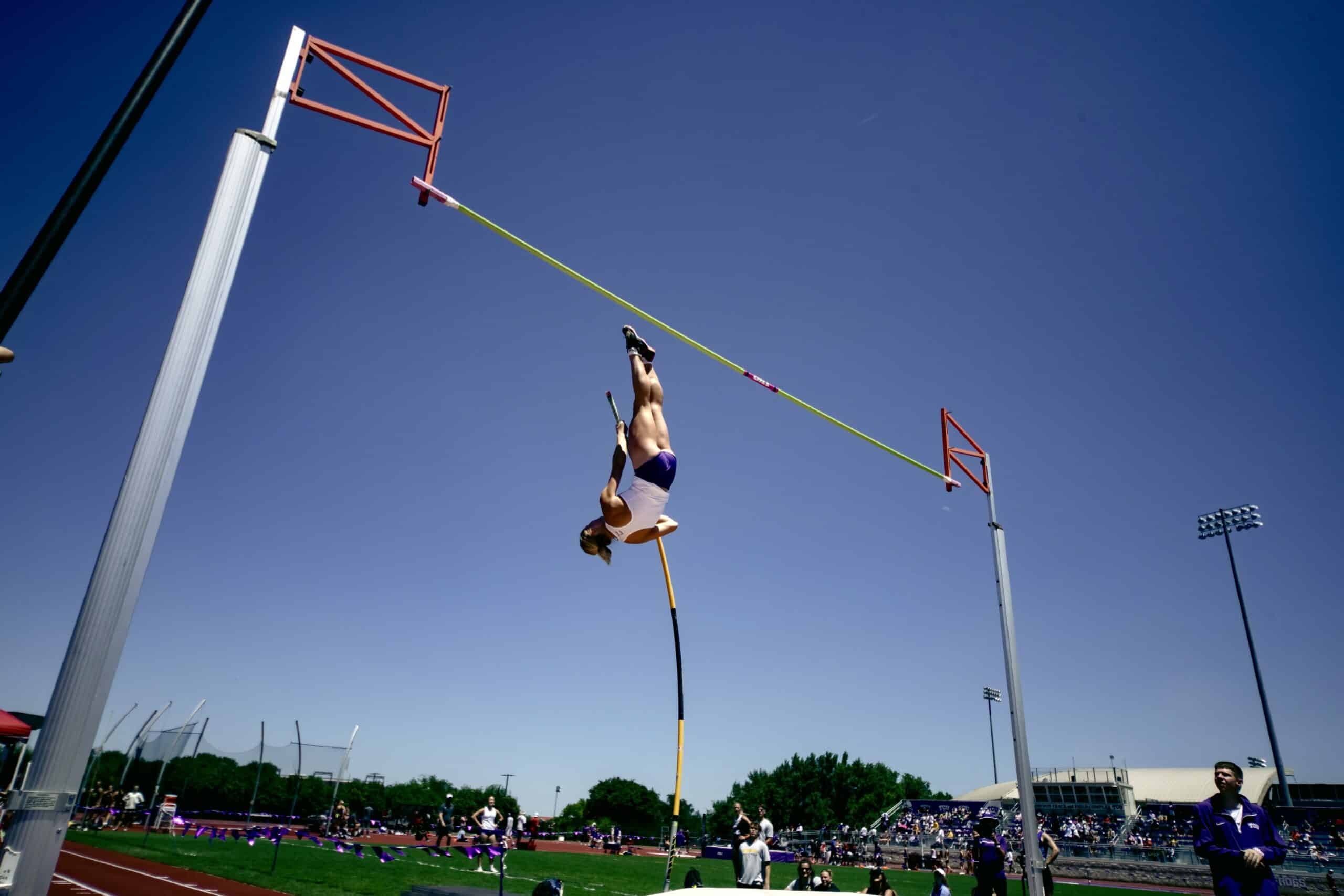 A pole vaulter is upside down in mid-air, holding a bent pole, attempting to clear a high bar on an outdoor athletics track under a clear blue sky. Spectators and athletes are visible in the background.