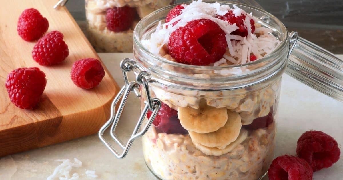 A glass jar filled with overnight oats, layered with banana slices, raspberries, and shredded coconut. Extra raspberries and coconut are scattered on a cutting board and marble surface nearby. Example breakfast option what to eat before track meet.