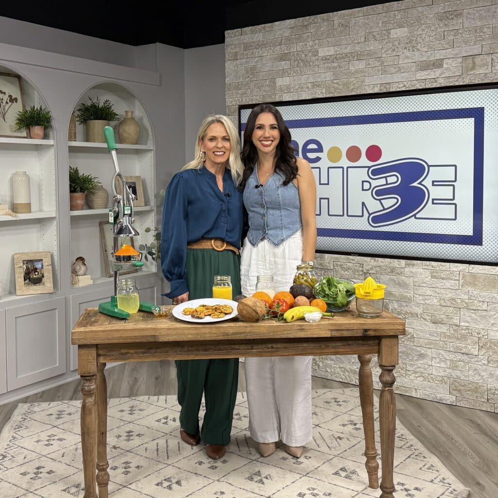 Jena Brown sports dietitian for endurance athletes and Lauren Margolis reporter stand smiling behind a wooden table with fruits, vegetables, a manual juicer, and plates of food. They are on a TV set with a sign reading The 3 on a brick wall in the background.