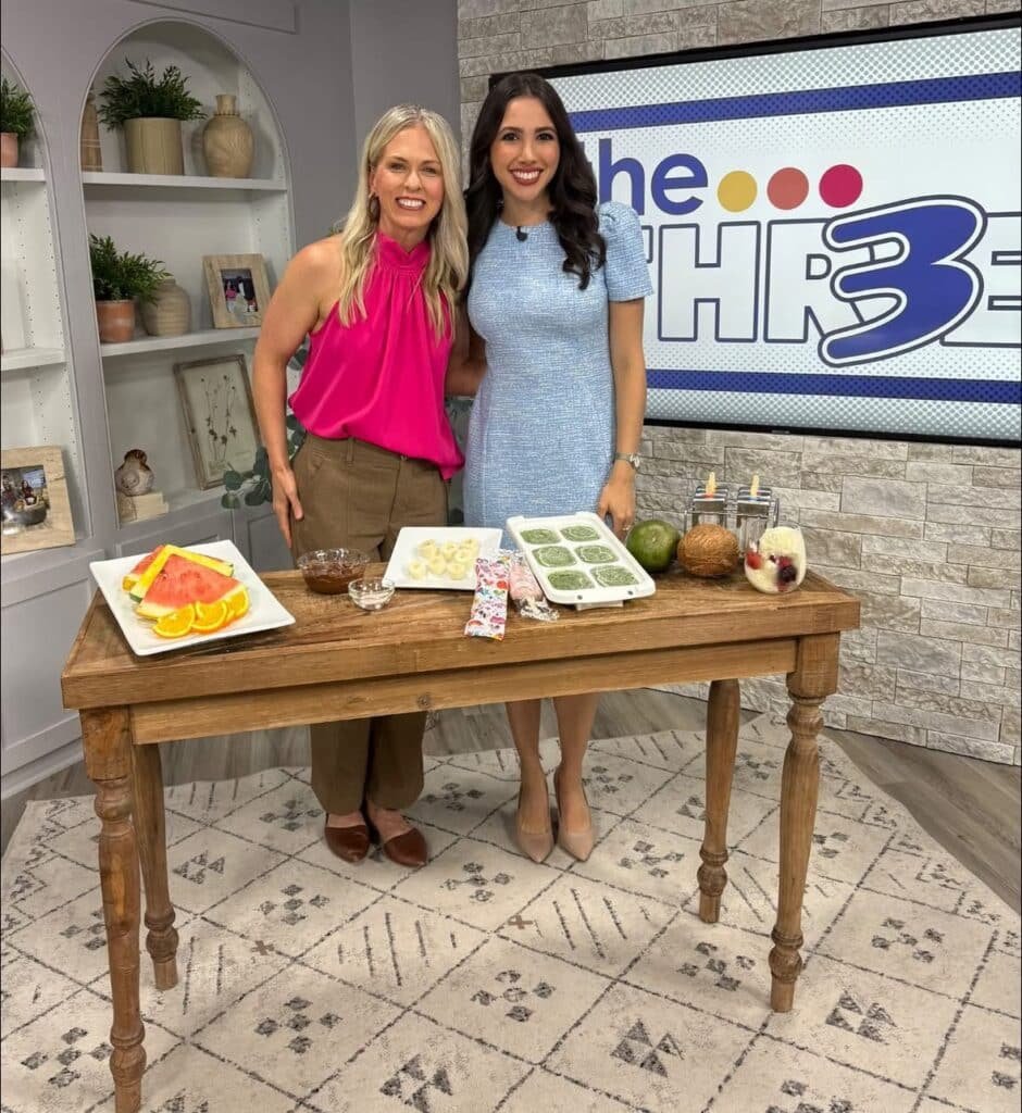 Jena Brown sports dietitian and Lauren Margolis reporter stand smiling behind a wooden table with fruit, smoothie packets, and ice cube trays, on the set of a TV show called The Three. Shelves and decor are visible in the background.