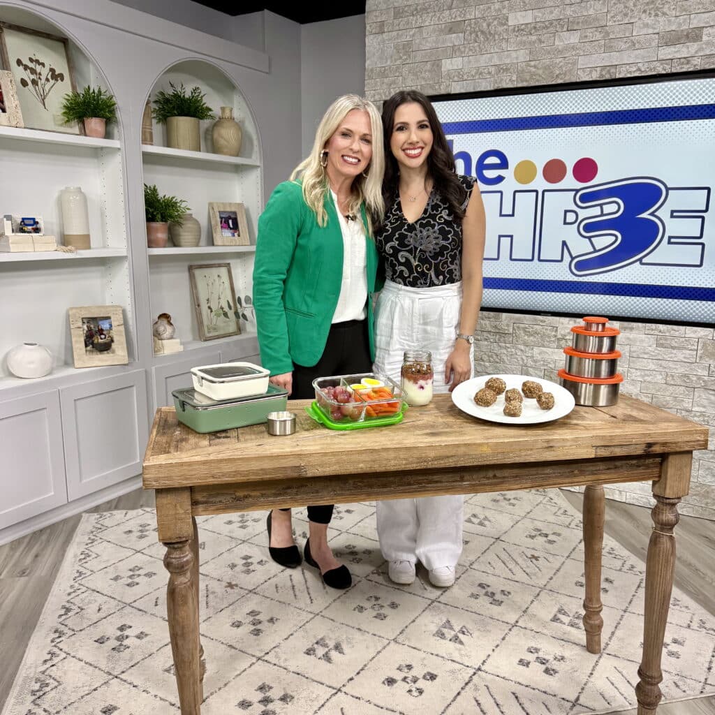 Jena Brown sports dietitian and Lauren Margolis reporter stand and smile behind a wooden table with snacks, including fruit, parfaits, and energy bites, in a bright studio. A The Three TV show sign is visible in the background.