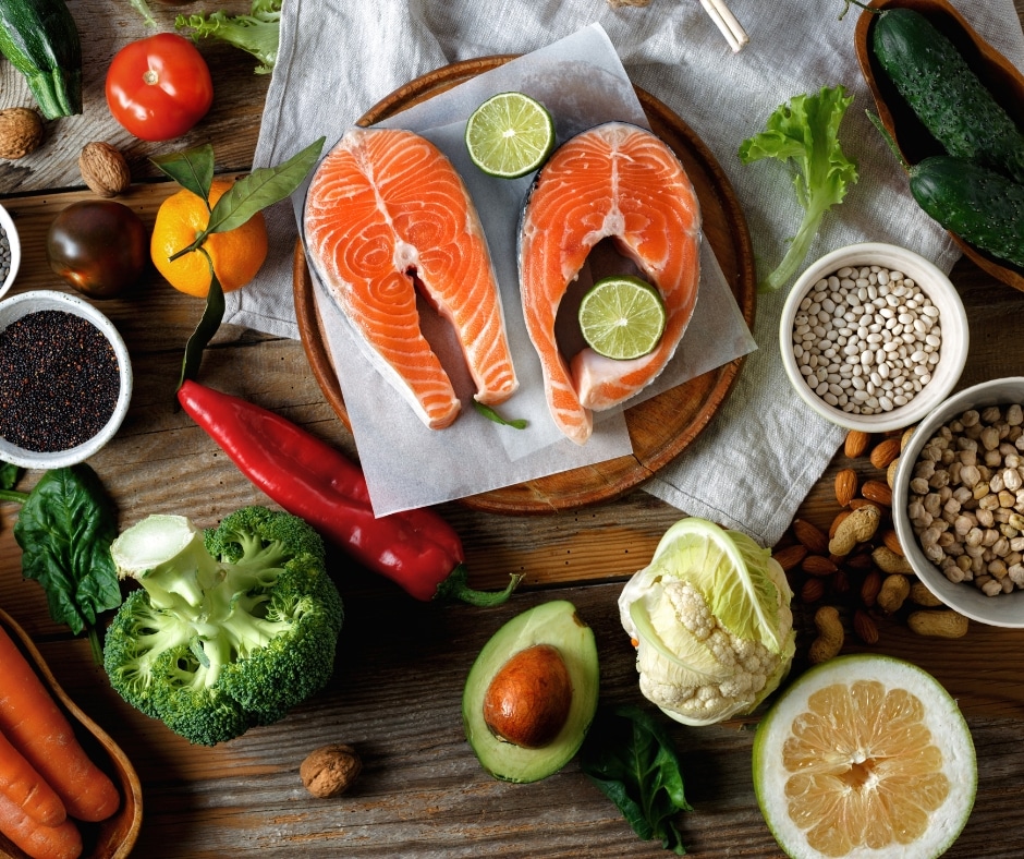 Flat lay out of nutrient-dense foods for adrenal health including salmon steaks, broccoli, avocado, carrots, citrus fruits, leafy greens, nuts, legumes, and quinoa on a rustic wooden table